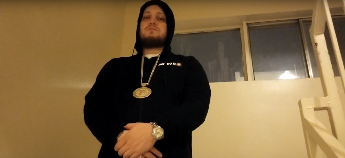Project Prezzy, wearing a black hoodie with a gold chain, stands looking at the camera in the new Work Music video.