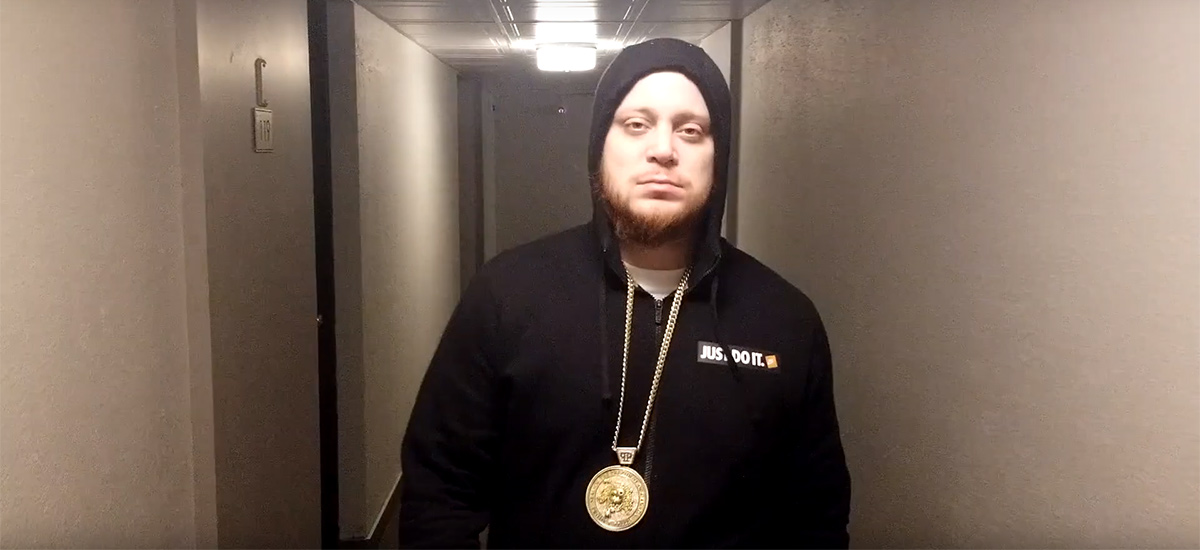 Project Prezzy, wearing a black hoodie with a gold chain, walks down the hall of an apartment building while looking at the camera in the new Work Music video.
