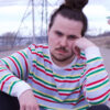 Nova scotian rapper Nomad Quinn in a red stripped white sweater, sitting on the road, resting his head against his hand.