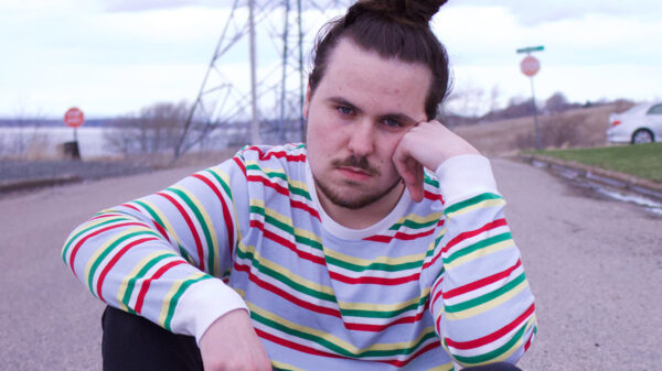 Nova scotian rapper Nomad Quinn in a red stripped white sweater, sitting on the road, resting his head against his hand.