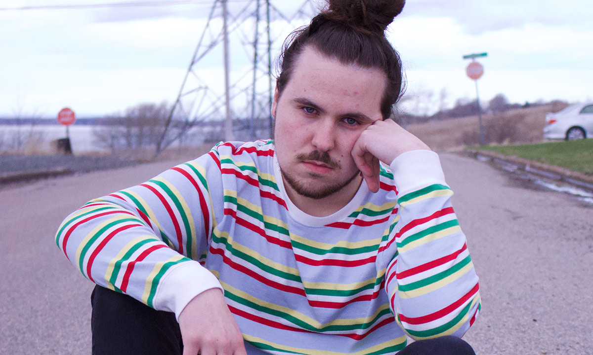Nova scotian rapper Nomad Quinn in a red stripped white sweater, sitting on the road, resting his head against his hand.