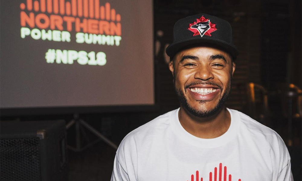 The image features Northern Power Summit organizer D.O. Gibson, smiling and standing in front of the a large screen with the summit logo on display.