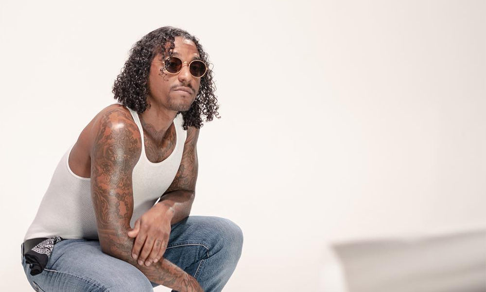 Photo of American singer Lloyd who is the subject of the interview in the post. He is crouched down in a white room, wearing blue jeans, a white tank top, and sunglasses.