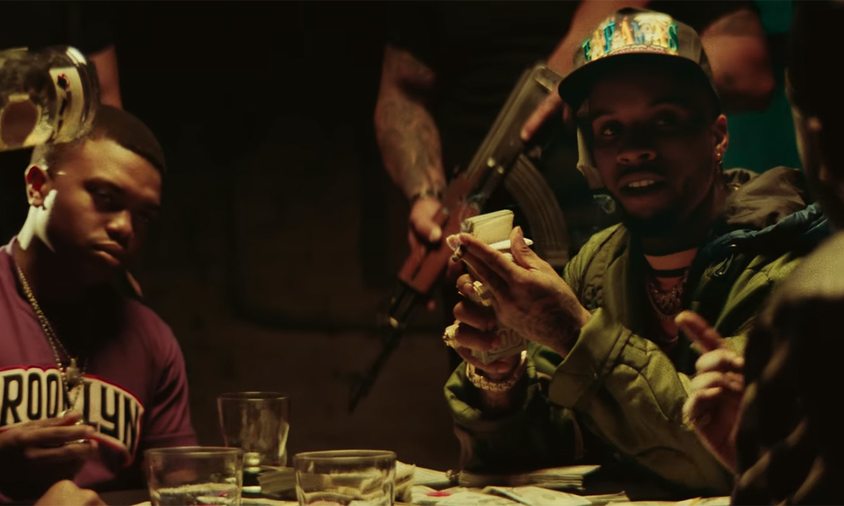 Tory Lanez is wearing a hat, seated at a table playing cards and holding a stack of money in his new video for Forever