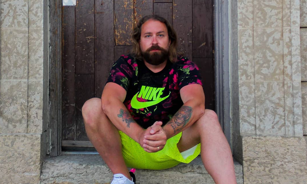Canadian rapper Fortunato sits on a stoop wearing a black Nike shirt with a fluorescent logo, and matching fluorescent coloured shorts.