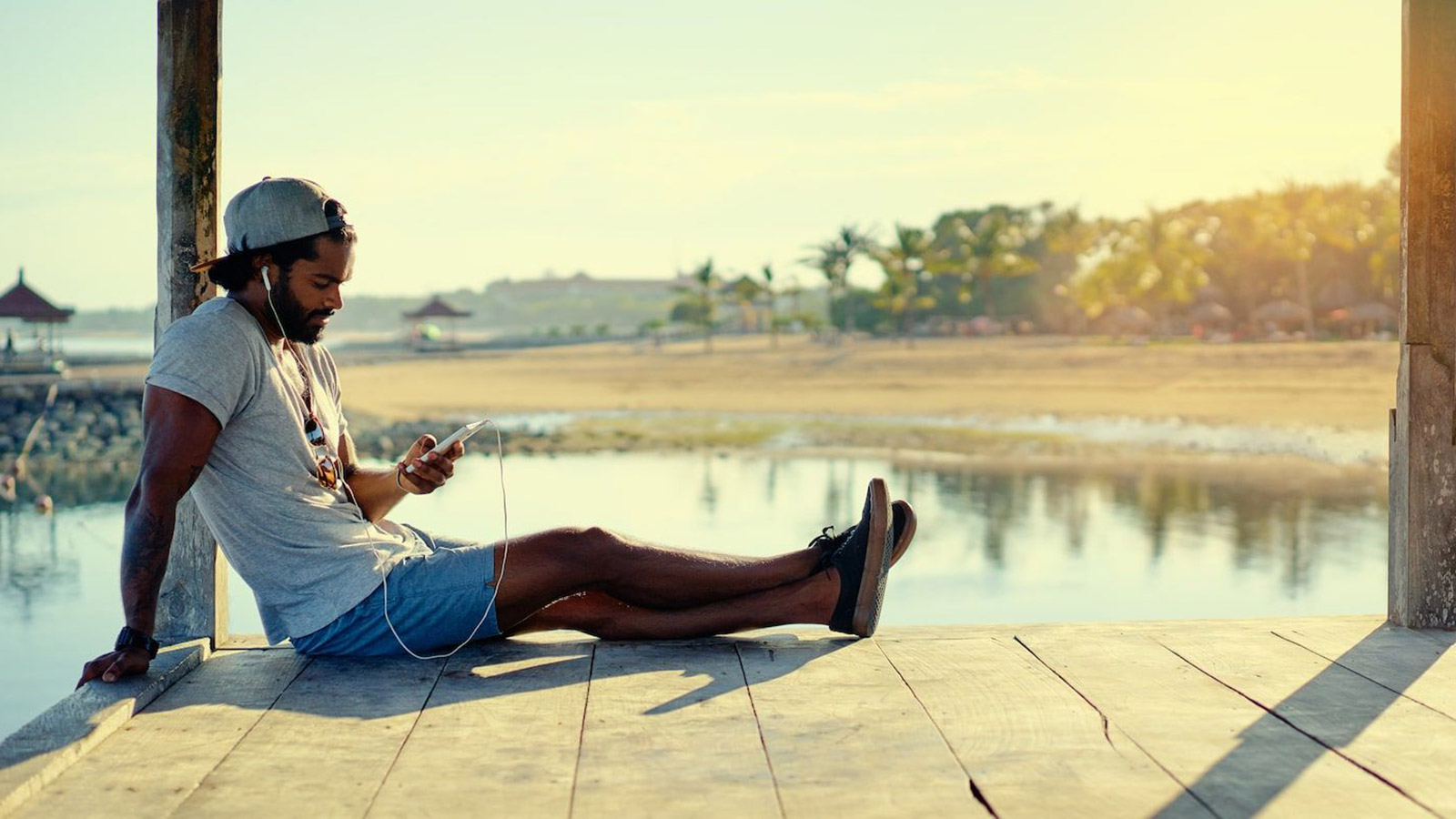 A man sits on a dock and looks at his phone with headphones on.