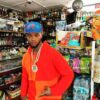Papoose in a convenience store wearing an orange and red hoodie and a blue hat