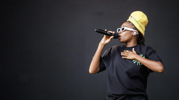 A woman wearing a yellow toque and black shirt holds a microphone to her mouth with her other hand on her chest.