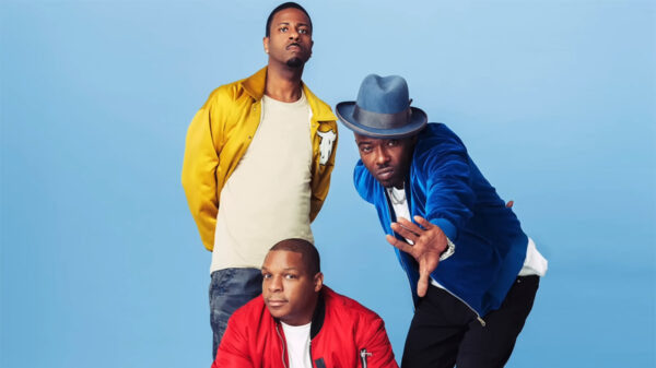 The 3-man rap group Naughty By Nature stands before a baby blue backdrop.