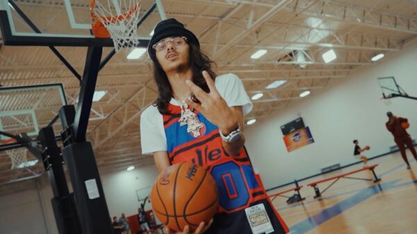 A man with glasses, a beanie and a basketball jersey holds a basketball on a basketball court.