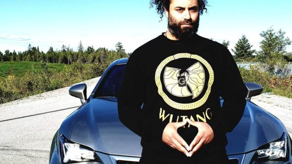 A man with curly hair in a black and gold Wu-Tang shirt sits on the hood of a car.