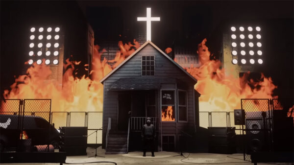 A man standing in front of a small church with a wall of fire in the background.