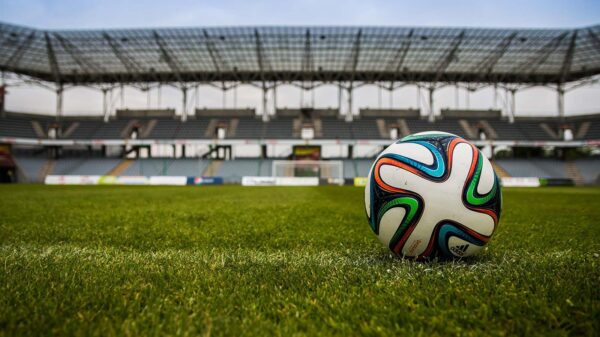A soccerball sits on a grass field in a large stadium during the daytime.