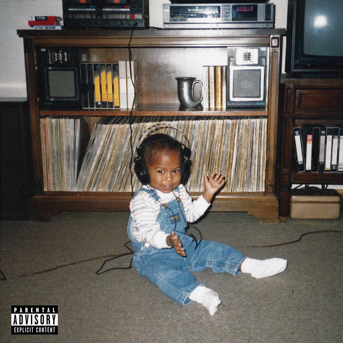 An older picture of a baby in overalls wearing headphones and sitting on the floor of a living room.