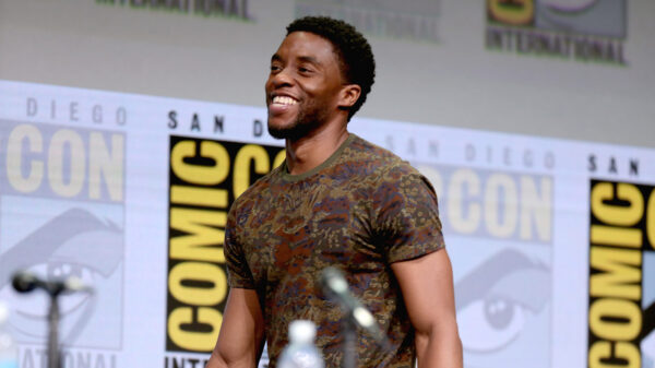 A Black man in a camoflauged shirt smiles while standing in front of a microphone and water bottle.