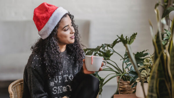 A woman wearing a Christmas sweater and Santa hat holds a cup of coffee.