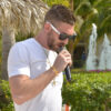 A man wearing a white t-shirt and sunglasses raps into a microphone outside on a sunny day