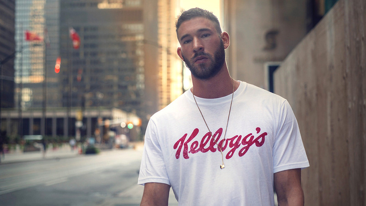 A man stands on a city street wearing a t-shirt with the word Kellong's written on it