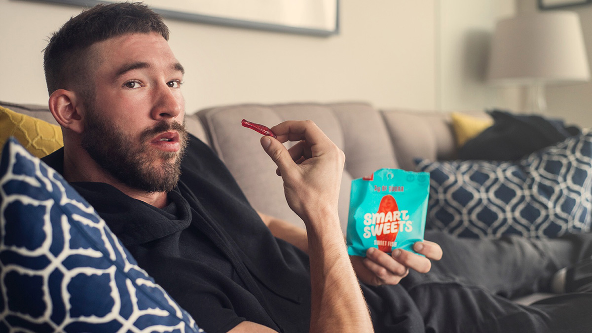 A man lounging on the couch while holding a bag of candy in one hand and a piece of candy in the other