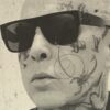 A man with a tattooed face wearing sunglasses, looking away from th camera