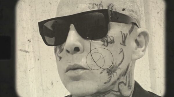 A man with a tattooed face wearing sunglasses, looking away from th camera