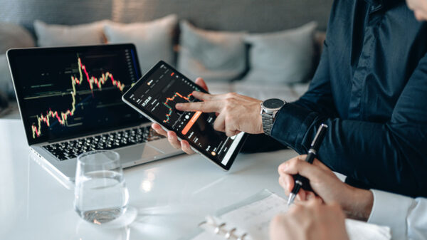 A hand holds a tablet and points to a stock market chart on the screen with the other.