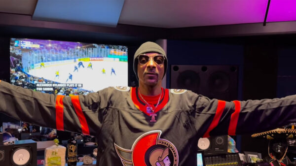A man wearing an Ottawa Senators hockey jersey extends his arms out while standing in a recording studio in front of a TV displaying a hockey game.