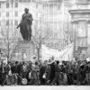 A large crowd protesting in front of a statue of a man.