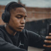 A young man sitting outside looks at a mobile device while wearing headphones.