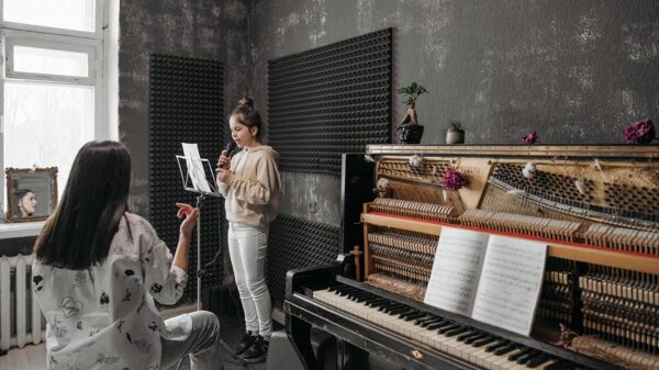 A girl reading a sheet music while singing next to a woman sitting at a piano.