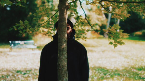 A man with a black hoodie and dark sunglasses stands behind a tree.