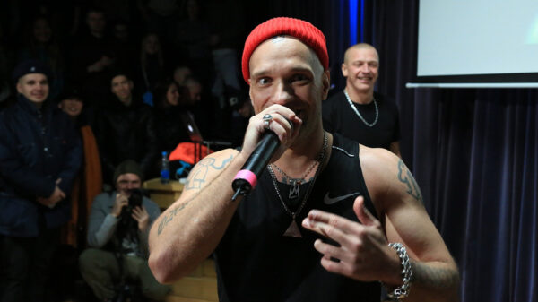A man with a red hat an a black tanktop holds a microphone to his face.
