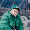 Rapper Peter Jackson wears a green hat and green jacket while sitting on bleechers in a scene from the Be Like Michael music video.