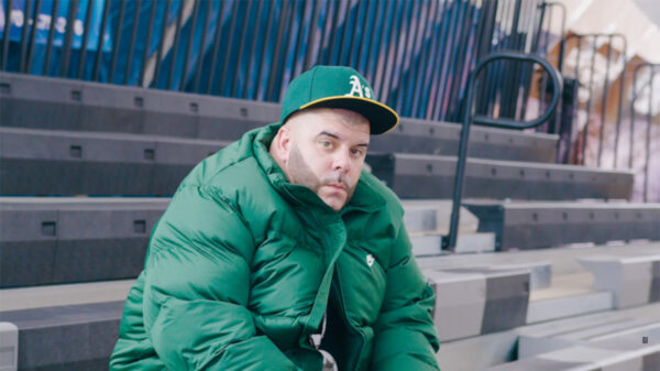 Rapper Peter Jackson wears a green hat and green jacket while sitting on bleechers in a scene from the Be Like Michael music video.