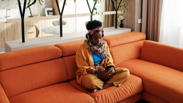 A woman sitting on an orange couch wears headphones while looking at a mobile phone.