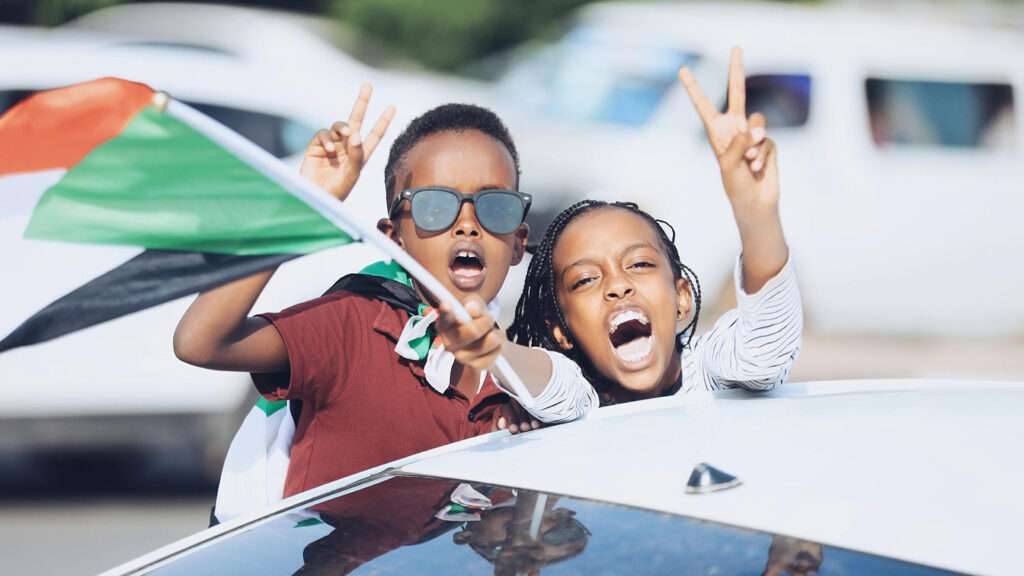 Two kids waving the flag of Sudan and throwing up peace signs.