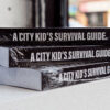 Three copies of 100: A City Kid's Survival Guide stacked on a table.