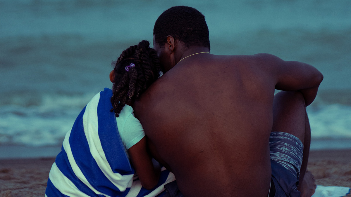 Two people sitting and leaning against each other facing a beach, on the poster for the film It Comes In Waves