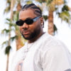 A recording artist, Shea Smilez, stands in front of palm trees while wearing a white t-shirt and sunglasses.