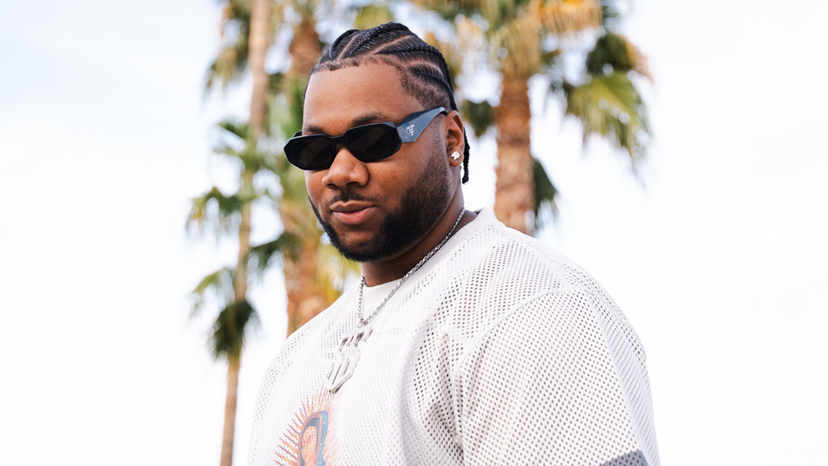 A recording artist, Shea Smilez, stands in front of palm trees while wearing a white t-shirt and sunglasses.
