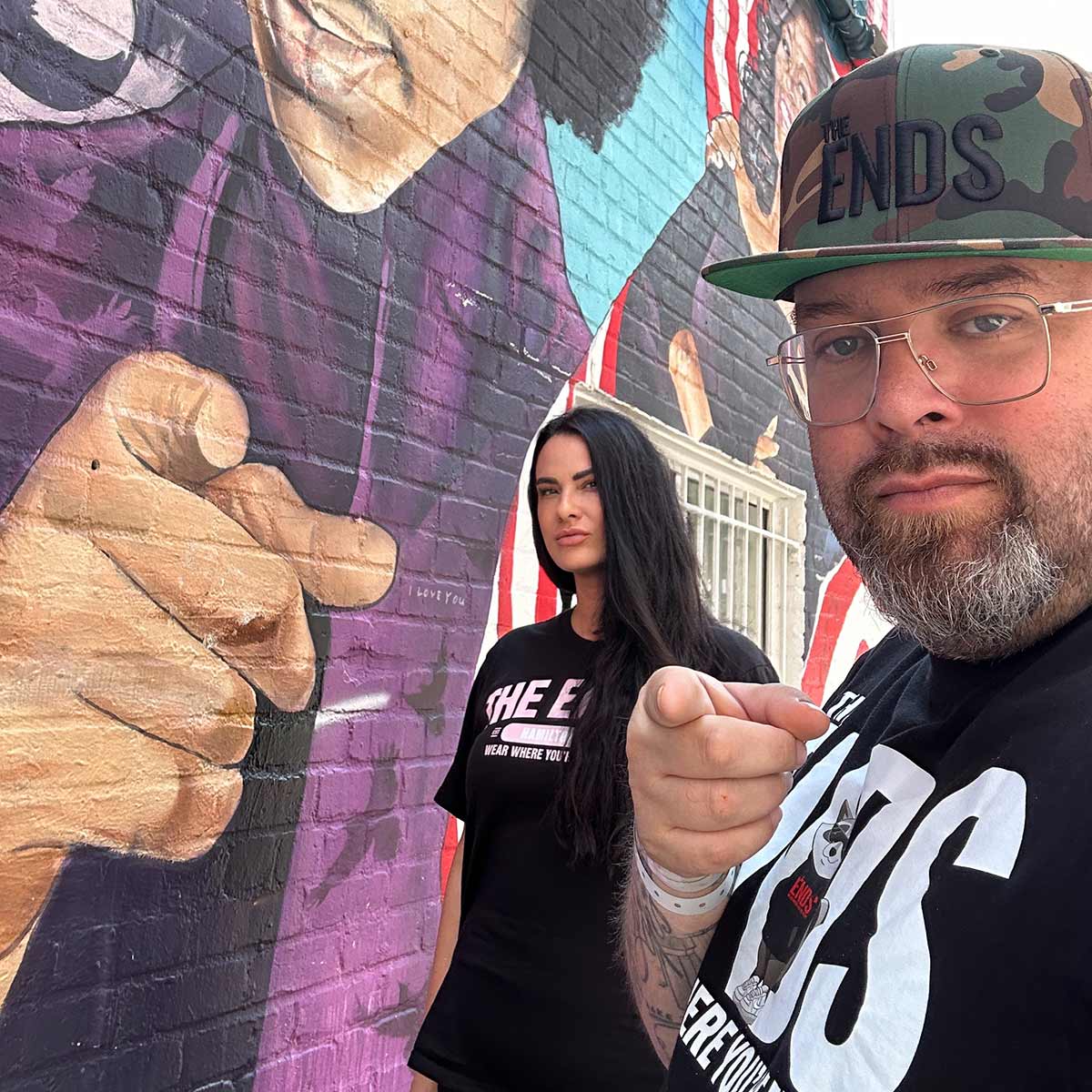 Melly poses for a photo with her partner Bishop Brigante next to a graffiti covered wall.