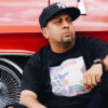 A man wearing a black shirt and a black NY Yankee fitted hat, leans against a red car.