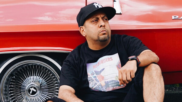 A man wearing a black shirt and a black NY Yankee fitted hat, leans against a red car.