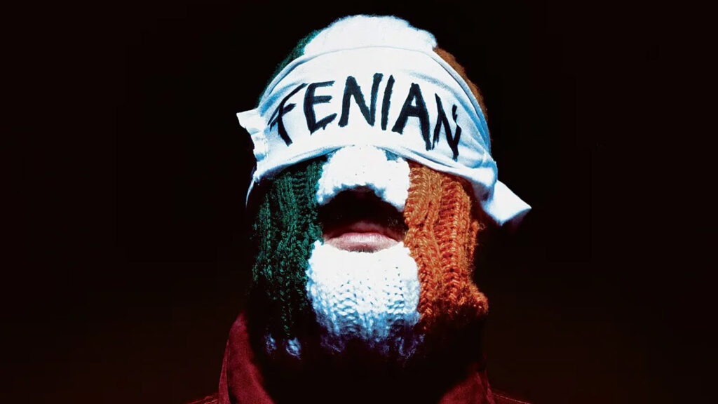 Kneecap member wearing a knitted tricolour, Irish-themed balaclava and a headband reading Fenian against a dark background.
