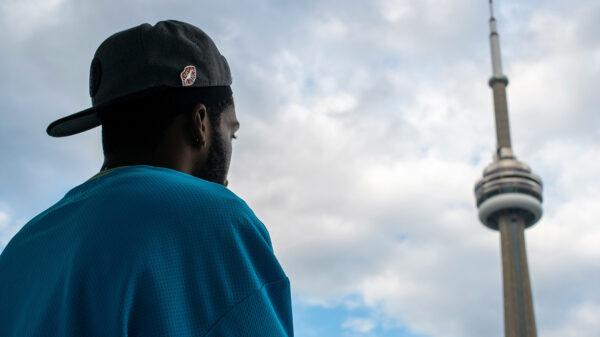 A young man wearing a baseball hat backwards looks up at the CN Tower in Toronto.