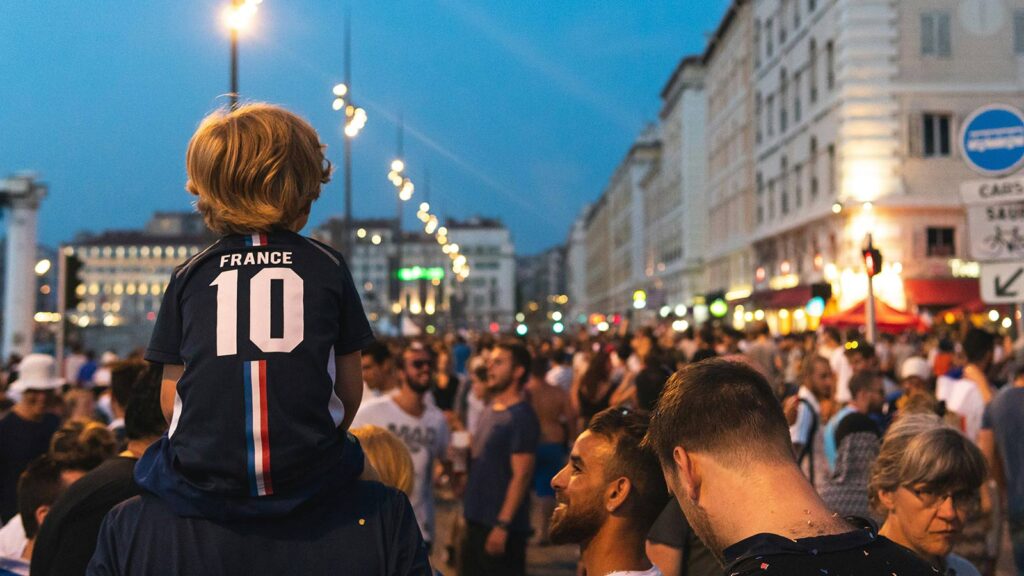 A young boy wearing a soccer jersey sits on an adults shoulders during a World Cup celebration outdoors.