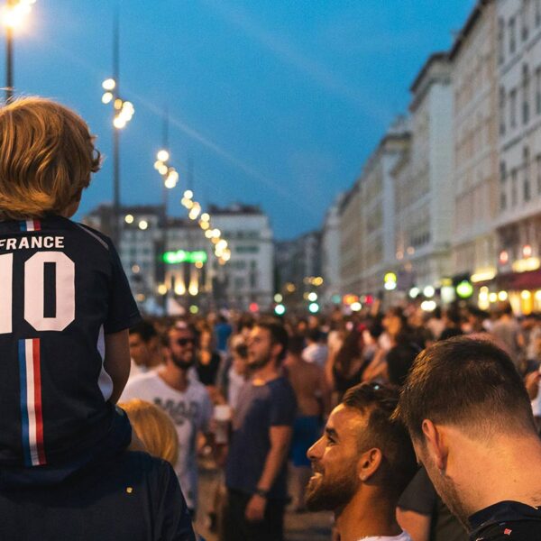 A young boy wearing a soccer jersey sits on an adults shoulders during a World Cup celebration outdoors.
